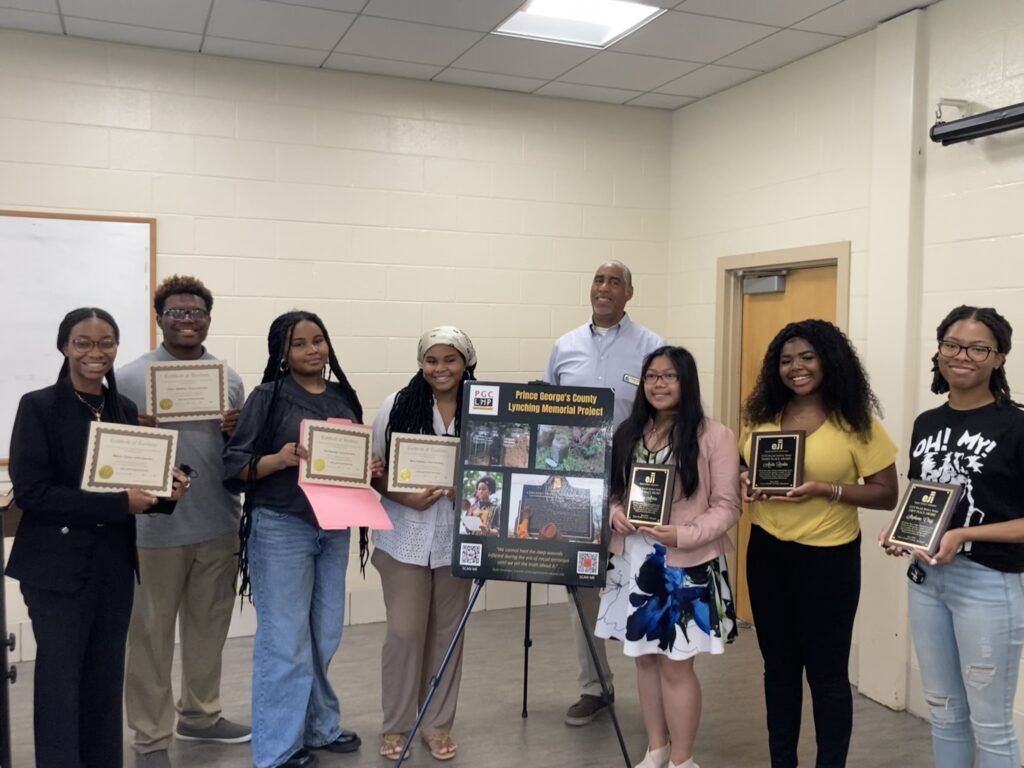 Students who won the essay and creative arts contest pose with award plaques and certificates around a posterboard for PGC LMP 